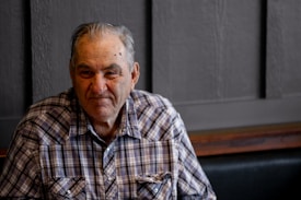 An elderly man with silver hair and a lined, weathered face sits in front of a dark paneled wall. He wears a plaid shirt and smiles subtly at the camera, conveying a sense of warmth and wisdom.