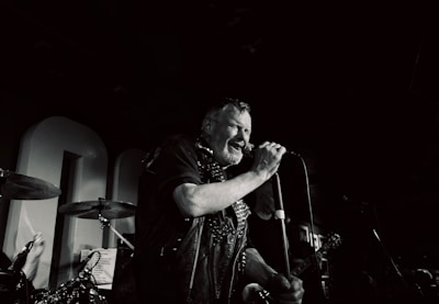 A middle-aged man passionately sings into a microphone on stage, surrounded by musical instruments. The setting suggests a live music performance in a dimly lit venue, with visible elements such as drums and a guitar.