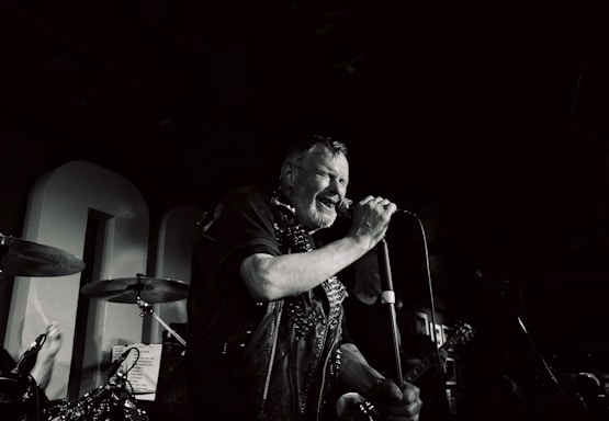 A middle-aged man passionately sings into a microphone on stage, surrounded by musical instruments. The setting suggests a live music performance in a dimly lit venue, with visible elements such as drums and a guitar.