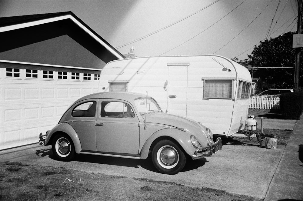 Foto en escala de grises del Volkswagen Beetle coupé junto a un remolque de viaje estacionado frente al garaje