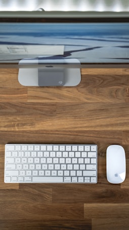 A minimalist desk setup featuring a wireless keyboard and mouse in white on a wooden surface. A computer monitor sits on a stand in the background, displaying part of a desktop screen with a landscape wallpaper.