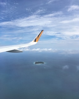 An airplane wing with a distinct yellow and black pattern is visible against a backdrop of a vast blue sky and ocean. Below, there's a small island surrounded by water, with scattered clouds enhancing the serene atmosphere.