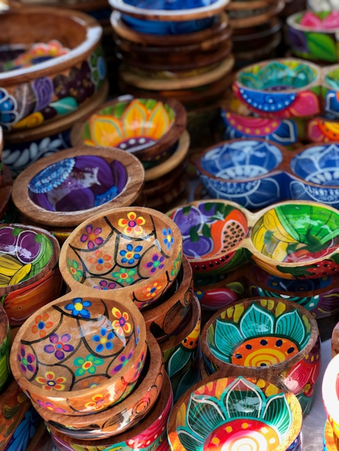 A collection of vibrant, hand-painted wooden bowls featuring various floral and geometric designs. Each bowl displays a unique pattern with bright colors such as blue, red, green, and yellow, arranged in a visually appealing manner. The collection is stacked together, showcasing the artistry and intricate details of the craftsmanship.
