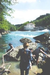 A group of people are exploring a rocky riverbank with clear blue water flowing and a building visible in the background surrounded by lush green trees. The sky is partly cloudy.