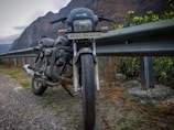 A motorcycle is parked on the side of a mountainous road, next to a metal guardrail. The road appears to be paved with gravel, and there are green shrubs and plants nearby. In the background, the towering mountains create a rugged landscape under a cloudy sky.