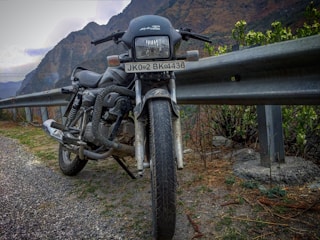 A motorcycle is parked on the side of a mountainous road, next to a metal guardrail. The road appears to be paved with gravel, and there are green shrubs and plants nearby. In the background, the towering mountains create a rugged landscape under a cloudy sky.