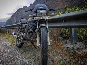 A motorcycle is parked on the side of a mountainous road, next to a metal guardrail. The road appears to be paved with gravel, and there are green shrubs and plants nearby. In the background, the towering mountains create a rugged landscape under a cloudy sky.