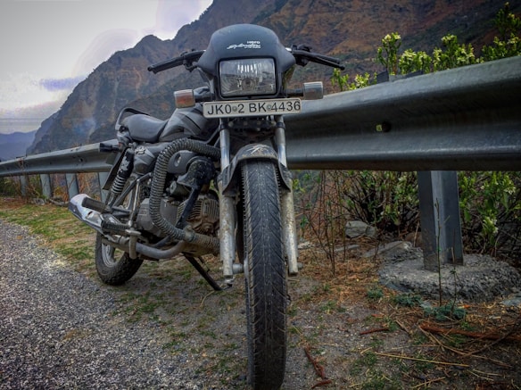A motorcycle is parked on the side of a mountainous road, next to a metal guardrail. The road appears to be paved with gravel, and there are green shrubs and plants nearby. In the background, the towering mountains create a rugged landscape under a cloudy sky.