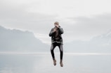 A person is captured mid-air while holding a camera up to their face, positioned against a serene backdrop of a misty lake and distant mountains under a cloudy sky. The scene conveys a sense of action and tranquility.