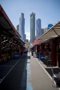 A bustling city market alley with stalls lining each side. The foreground shows a seating area with tables and chairs, and further down, people are walking and browsing. Towering skyscrapers with cranes are visible against a clear blue sky in the background.