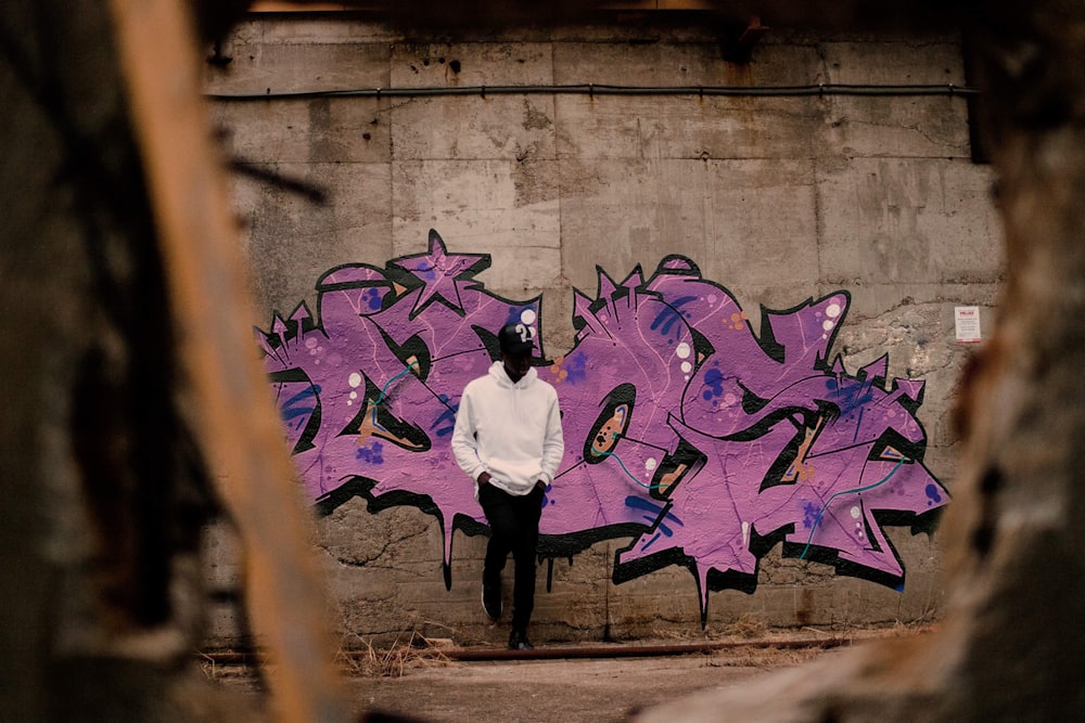 homme en pull à capuche blanc s’appuyant sur un mur violet tagué