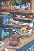 A display of baked goods in a café, featuring a chocolate loaf cake on a wooden board and a slice of green cake garnished with strawberries on a glass stand. Behind them are various kitchen items, a coffee machine, and shelves stocked with different products. The words 'Pretty Cuppa' are visible on the glass.