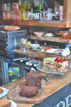 A display of baked goods in a café, featuring a chocolate loaf cake on a wooden board and a slice of green cake garnished with strawberries on a glass stand. Behind them are various kitchen items, a coffee machine, and shelves stocked with different products. The words 'Pretty Cuppa' are visible on the glass.