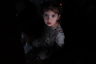 A young child with wide eyes and curly hair looks directly at the camera. She is wearing a floral patterned dress. The lighting is dim, creating a dramatic contrast, with her face and dress softly illuminated.