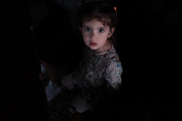 A young child with wide eyes and curly hair looks directly at the camera. She is wearing a floral patterned dress. The lighting is dim, creating a dramatic contrast, with her face and dress softly illuminated.