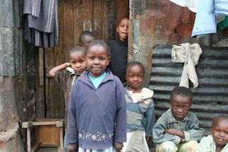 A group of children standing in front of a makeshift structure made from wood and corrugated metal. They are smiling and wearing casual clothing. Some clothes are hanging on the wall outside the structure.
