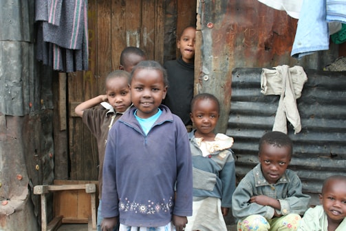 A group of children standing in front of a makeshift structure made from wood and corrugated metal. They are smiling and wearing casual clothing. Some clothes are hanging on the wall outside the structure.