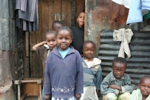 A group of children standing in front of a makeshift structure made from wood and corrugated metal. They are smiling and wearing casual clothing. Some clothes are hanging on the wall outside the structure.