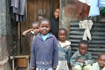 A group of children standing in front of a makeshift structure made from wood and corrugated metal. They are smiling and wearing casual clothing. Some clothes are hanging on the wall outside the structure.