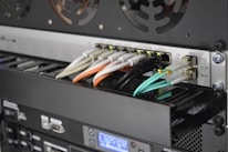 A close-up view of the back of a server rack with multiple network cables connected to a switch. The cables are in different colors including white, orange, black, and light blue. There are also various ports, indicators, and a digital display indicating some operational parameters.