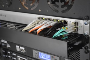 A close-up view of the back of a server rack with multiple network cables connected to a switch. The cables are in different colors including white, orange, black, and light blue. There are also various ports, indicators, and a digital display indicating some operational parameters.