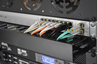A close-up view of the back of a server rack with multiple network cables connected to a switch. The cables are in different colors including white, orange, black, and light blue. There are also various ports, indicators, and a digital display indicating some operational parameters.