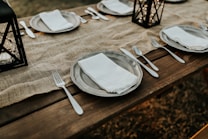 A rustic wooden table set for a meal, featuring plates with white napkins and silverware neatly arranged. Burlap serves as a table runner, and decorative lanterns are placed as centerpieces. The setting is outdoors, creating a cozy and inviting ambiance.