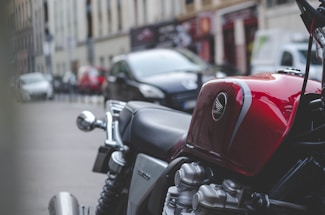 A close-up of a classic red motorcycle with a shiny gas tank featuring the Honda logo. The background shows a blurred urban street with cars parked along the road and buildings lining the street.