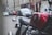 A close-up of a classic red motorcycle with a shiny gas tank featuring the Honda logo. The background shows a blurred urban street with cars parked along the road and buildings lining the street.