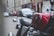 A close-up of a classic red motorcycle with a shiny gas tank featuring the Honda logo. The background shows a blurred urban street with cars parked along the road and buildings lining the street.