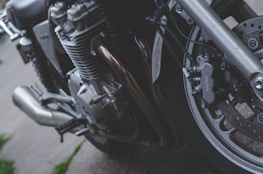A close-up view of a motorcycle's engine and front wheel. The image highlights the metal parts, including the brake caliper labeled 'Nissin', exhaust pipes, and complex engine components. The setting appears to be outdoor on a pavement.