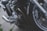 A close-up view of a motorcycle's engine and front wheel. The image highlights the metal parts, including the brake caliper labeled 'Nissin', exhaust pipes, and complex engine components. The setting appears to be outdoor on a pavement.