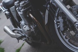 A close-up view of a motorcycle's engine and front wheel. The image highlights the metal parts, including the brake caliper labeled 'Nissin', exhaust pipes, and complex engine components. The setting appears to be outdoor on a pavement.