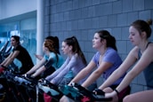 A group of people are exercising on stationary bicycles indoors. They are wearing casual workout clothes, some with earbuds and armbands, suggesting they might be listening to music or a workout program. The background features a tiled wall and glass windows, giving the scene a modern gym atmosphere.