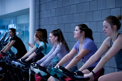 A group of people are exercising on stationary bicycles indoors. They are wearing casual workout clothes, some with earbuds and armbands, suggesting they might be listening to music or a workout program. The background features a tiled wall and glass windows, giving the scene a modern gym atmosphere.