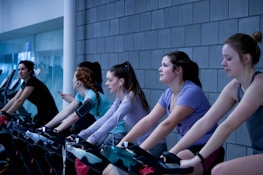 A group of people are exercising on stationary bicycles indoors. They are wearing casual workout clothes, some with earbuds and armbands, suggesting they might be listening to music or a workout program. The background features a tiled wall and glass windows, giving the scene a modern gym atmosphere.