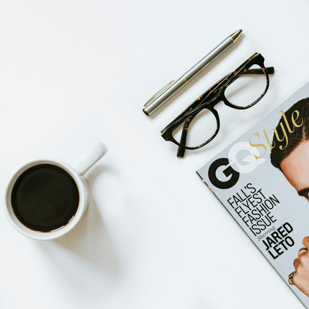 klare Brille mit schwarzem Rahmen in der Nähe einer weißen Keramik-Kaffeetasse