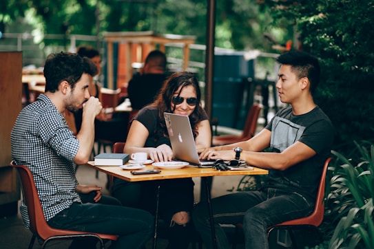 Three people are sitting at an outdoor table engaged in conversation. There is a laptop on the table, along with cups and other small items. They appear to be enjoying a casual discussion or collaborative work session. The background includes greenery and other seated individuals.
