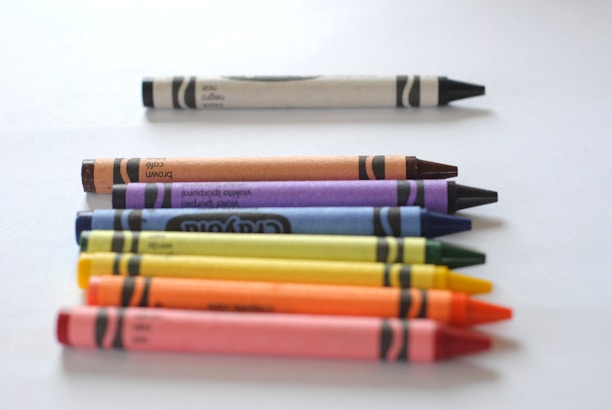 A collection of eight crayons is neatly arranged on a white surface in a diagonal line. A single white crayon is positioned horizontally at the top, separate from the rest. The other crayons below it, arranged from left to right, are brown, purple, blue, green, yellow, orange, and red. They create a vibrant and colorful display.