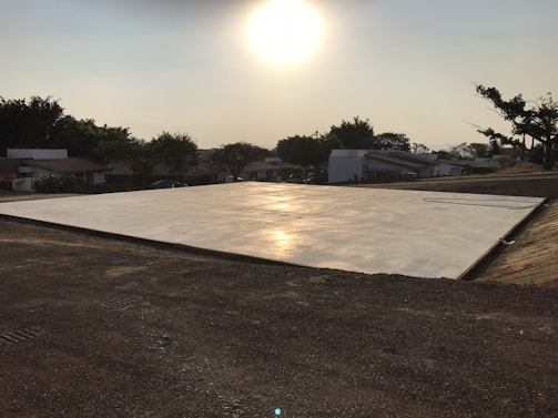 A flat concrete slab is illuminated by the setting sun, casting a warm glow across its surface. Surrounding the slab is an area of dirt and some sparse trees and houses in the distance under a clear sky.