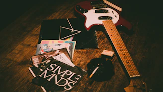 An electric guitar with a red body and wooden neck lies on a wooden surface alongside several vinyl records, some featuring colorful and bold designs. Nearby, a pack of cigarettes and an ashtray with a cigarette add to the scene, creating a vintage, rock-and-roll ambiance.