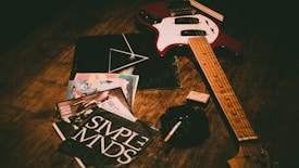 An electric guitar with a red body and wooden neck lies on a wooden surface alongside several vinyl records, some featuring colorful and bold designs. Nearby, a pack of cigarettes and an ashtray with a cigarette add to the scene, creating a vintage, rock-and-roll ambiance.