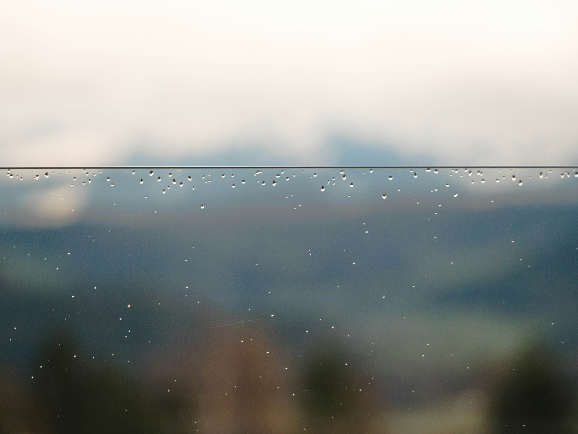 A close-up of raindrops splashing on a windowpane, with blurred green trees outside.