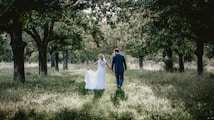 A couple walks hand in hand through a serene forest path. The bride wears an elegant white dress, and the groom is in a blue suit. Sunlight filters through the canopy of lush green leaves, casting soft shadows on the grassy ground.
