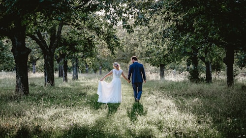 A couple walks hand in hand through a serene forest path. The bride wears an elegant white dress, and the groom is in a blue suit. Sunlight filters through the canopy of lush green leaves, casting soft shadows on the grassy ground.