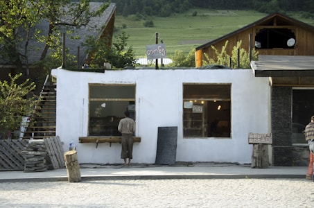 A small, rustic building with a white facade and two large windows. One person is standing in front of the building, possibly looking inside. There is a sign reading 'PURI' in one window. The area is surrounded by greenery, and the building has a wooden roof. Nearby, there are tree logs and a makeshift fence made of wooden pallets. Another person stands to the right, partially visible.