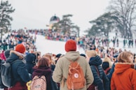 A large crowd of people gathered on a snowy slope, with many wearing colorful winter clothing. Trees frame the scene, and a small building is visible in the background. The crowd seems to be participating in an outdoor event or festival.