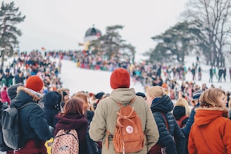 A large crowd of people gathered on a snowy slope, with many wearing colorful winter clothing. Trees frame the scene, and a small building is visible in the background. The crowd seems to be participating in an outdoor event or festival.