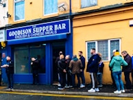 People are standing in line outside a supper bar named 'The Goodison Supper Bar' which advertises English and Chinese meals. The exterior of the building is painted yellow and blue. There are around 10 individuals, some interacting and some waiting in line. One person is holding food on a tray. The mood suggests a casual and somewhat social environment.