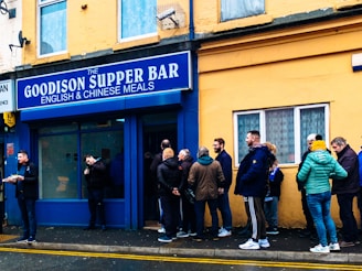 People are standing in line outside a supper bar named 'The Goodison Supper Bar' which advertises English and Chinese meals. The exterior of the building is painted yellow and blue. There are around 10 individuals, some interacting and some waiting in line. One person is holding food on a tray. The mood suggests a casual and somewhat social environment.